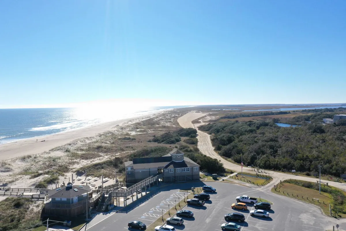 An expansive view of the beach and ocean, with a wooden boardwalk crossing the dunes. The scene also includes a parking area for easy access and open sandy spaces ideal for sunbathing, beach games, or simply enjoying the seascape.