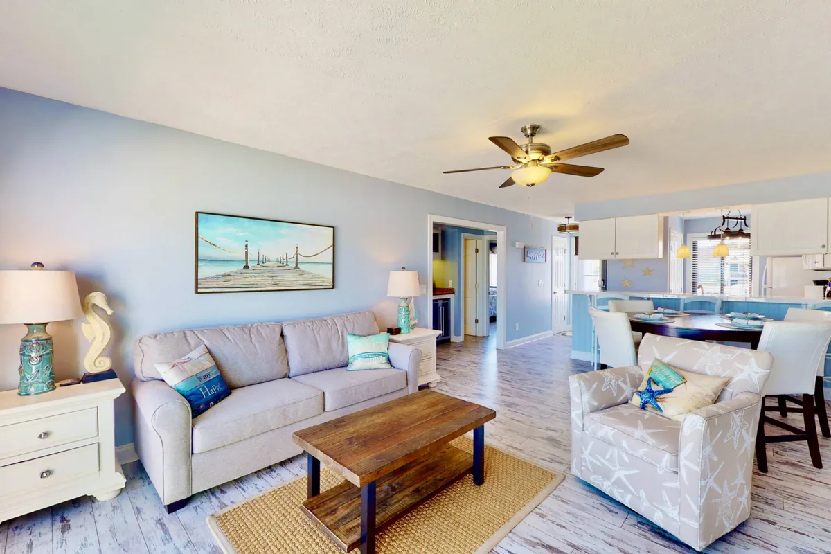 A comfortable living area with a stylish sofa, wooden coffee table, art on the wall, ceiling fan, and glimpse of a dining area. The space is arranged for relaxation and socializing, hinting at its versatility as both a gathering spot and an additional sleeping area.