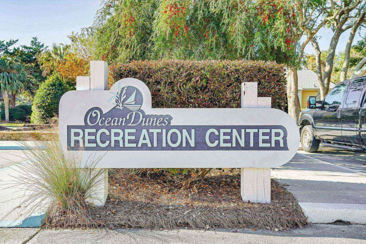 The exterior of a recreation center is presented, marked by a clear sign and surrounded by greenery. Vehicles parked nearby hint at community accessibility and the welcoming nature of the communal spaces, such as courts, playgrounds, and event areas.