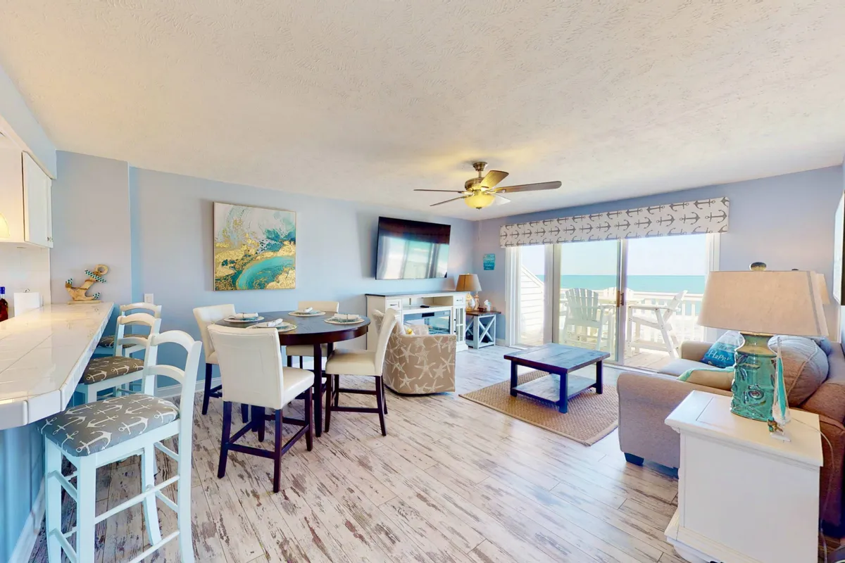 A spacious, open-concept living area featuring a comfortable sofa, a dining table with bar seating, and a large window that frames an expansive ocean view. The space is accented by a modern Smart TV and contemporary decor, providing a bright and welcoming atmosphere.