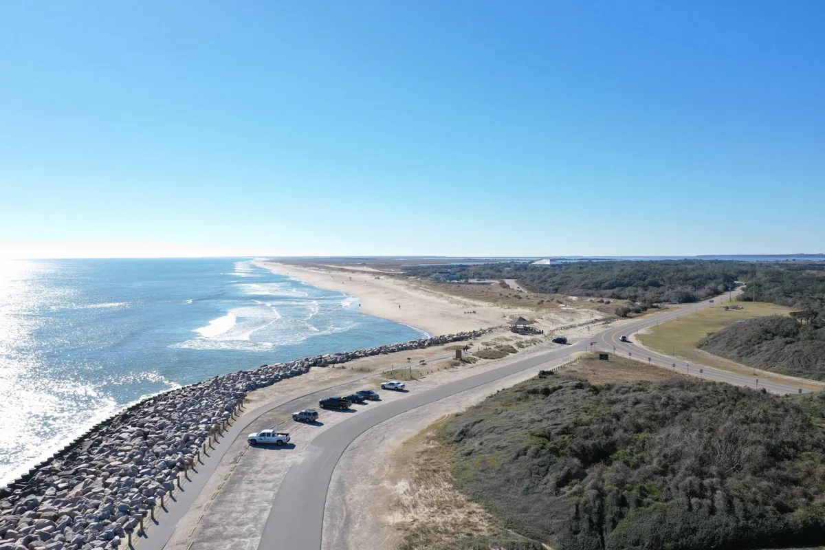 A panoramic view of the beach and ocean with dunes, a jetty in the distance, and people engaging in various activities along the shore. Vehicles are visible, suggesting convenient beach access for carrying equipment for water sports or picnics.