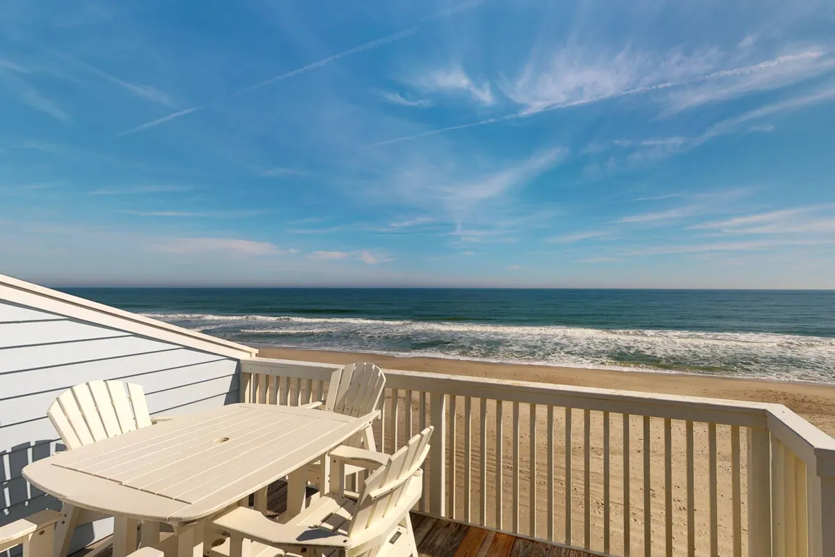 The hero image showcases a top-floor balcony with comfortable patio furniture overlooking endless ocean views and a pristine sandy beach. Gentle sunlight bathes the deck, suggesting the perfect spot for morning coffee or evening sunset dining. The serene atmosphere invites relaxation, with the soothing sound of waves and a cool ocean breeze adding to the allure.