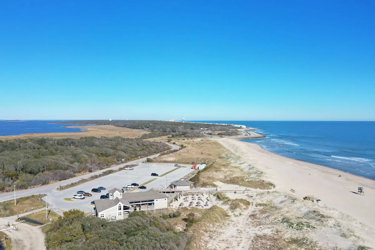 This view captures the beach and ocean alongside a convenient parking area and a nearby restaurant building. The scene combines leisure and practicality, suggesting easy access to dining, amenities, and waterside relaxation.