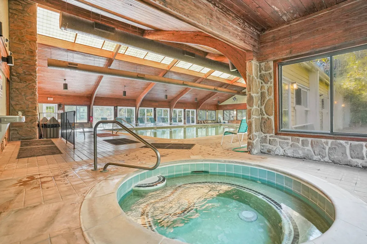 An inviting indoor pool is the main feature, with a bubbling hot tub adjacent to it. Large windows flood the space with light, while wooden beams add warmth. The scene is set for both swimming year-round and unwinding in the comfort of the hot tub, regardless of the season.