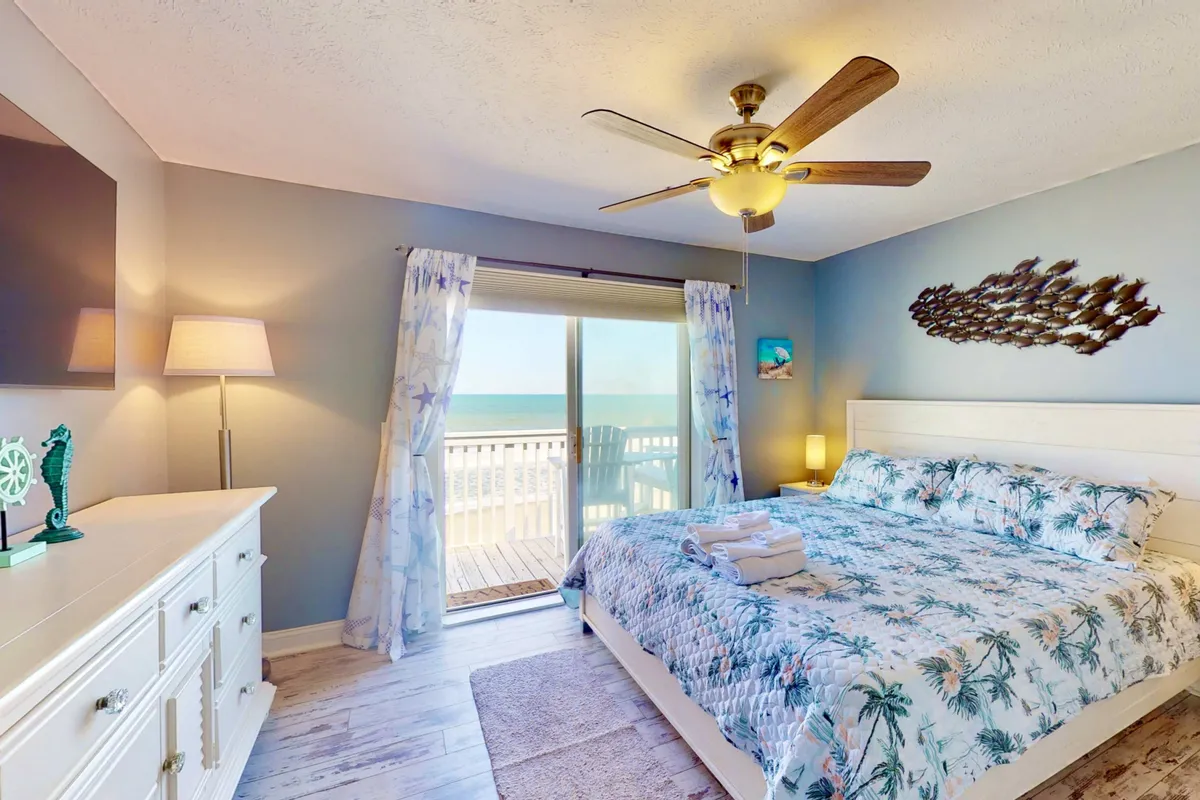 A tranquil master bedroom featuring a bed with tropical-themed bedding, a large window offering an ocean view, decorative fish wall art, and a cozy dresser with a lamp. The space is bathed in natural light and exudes a serene, inviting ambiance perfect for rest.