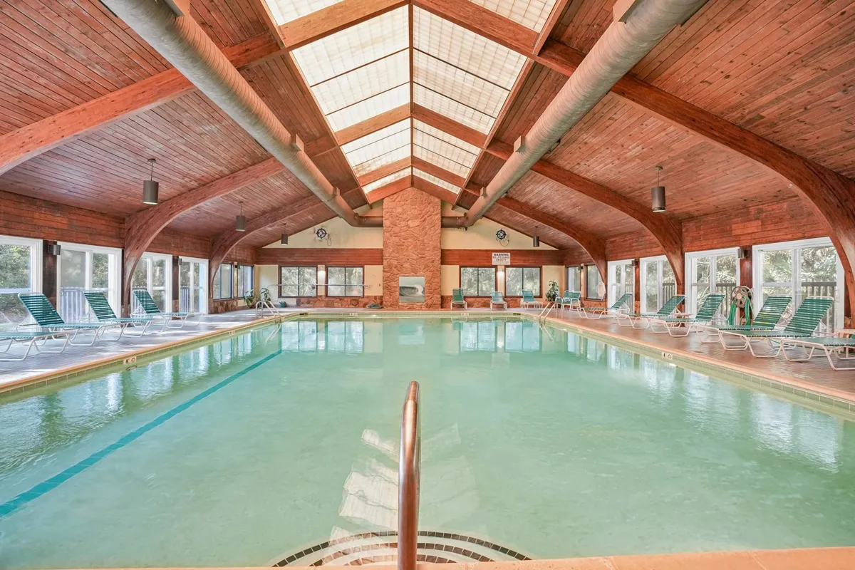 This image showcases the large indoor heated pool area, complete with wooden beams, expansive windows letting in sunlight, and several lounge chairs. The tranquil setting is ideal for swimming, relaxing, and socializing, regardless of the weather.