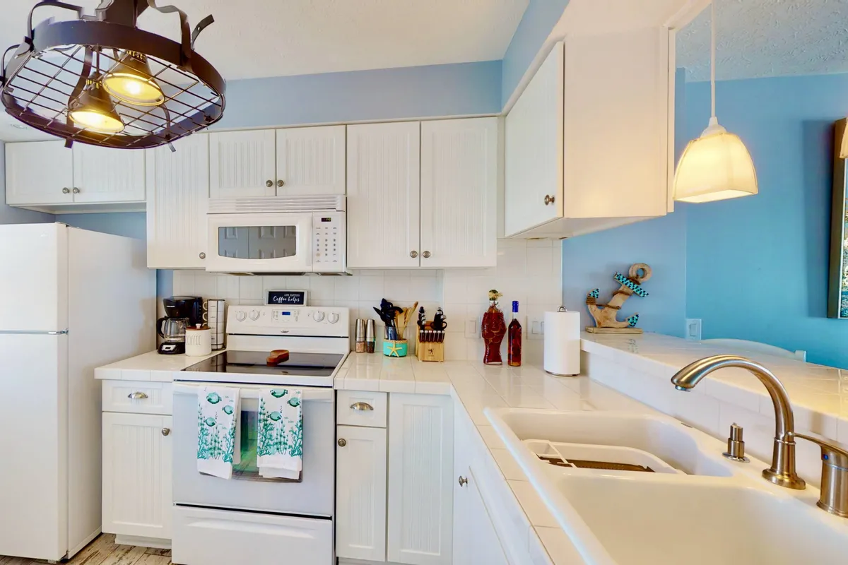 A bright, modern kitchen featuring sleek white cabinetry, stainless steel appliances, colorful dish towels, and a unique light fixture. Countertops are uncluttered, showcasing ample workspace and inviting cooking ambiance.