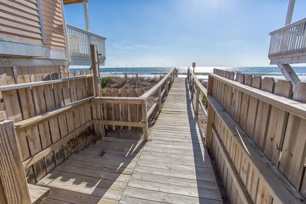 This scenic exterior image features a wooden walkway leading across dune grass directly to the wide, inviting beach and the rolling ocean beyond. It encapsulates the ease of access to sandy shores and the natural beauty of the surrounding area.